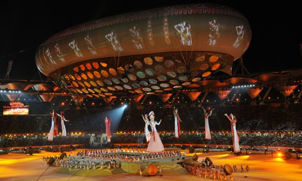 A grand stadium is filled with vibrant lights and a large audience. The scene features an elaborate stage with numerous performers in traditional costumes. Towering above the stage, several large, colourful puppets or figures are suspended in the air, giving a sense of grandeur and festivity. The background includes a massive overhead structure adorned with intricate patterns and illuminated by projected images of dancers. The atmosphere is lively and celebratory, suggesting a significant cultural or ceremonial event.