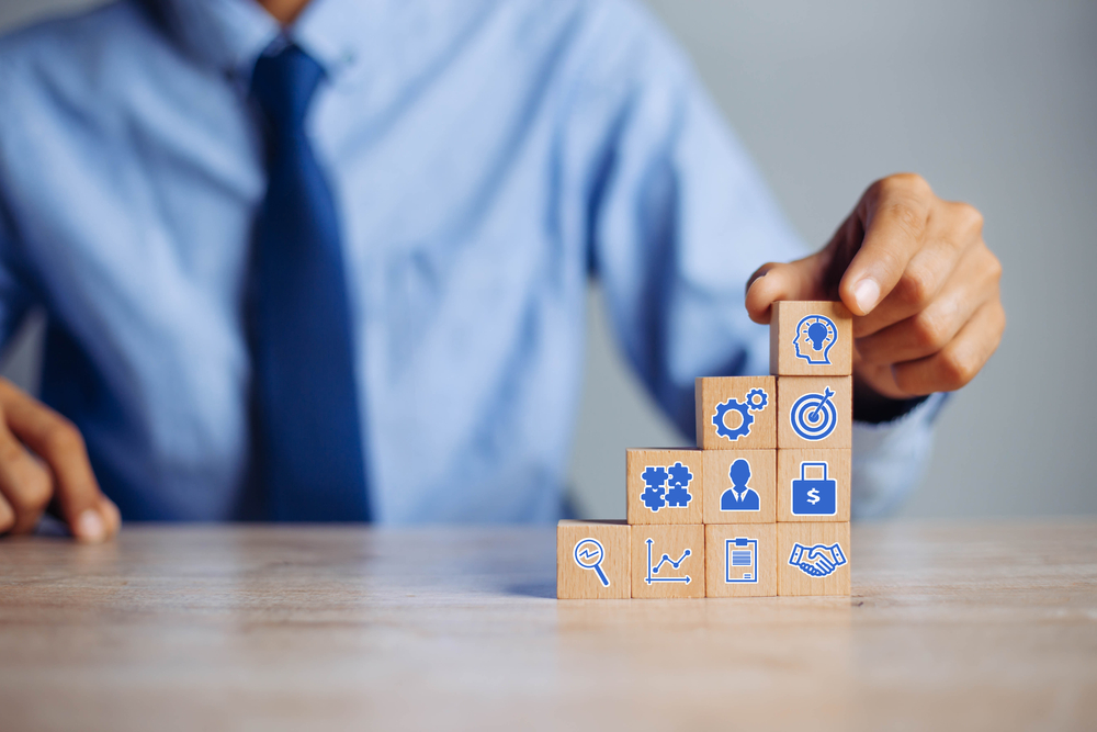 Close-up of a person in business attire stacking wooden blocks with blue icons representing key elements of strategic planning and goal achievement, including innovation, teamwork, financial growth, and leadership.