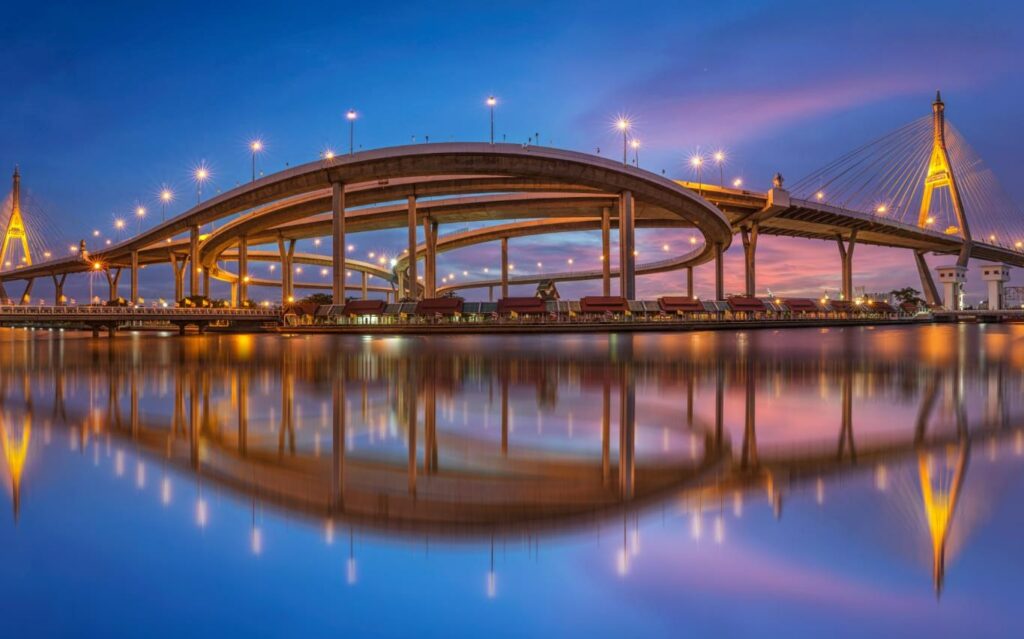 A large illuminated suspension bridge with curving roadways stretches across a body of water at dusk. The bridge’s lights and structure create a vivid reflection on the calm surface of the water, blending with the soft hues of the twilight sky.