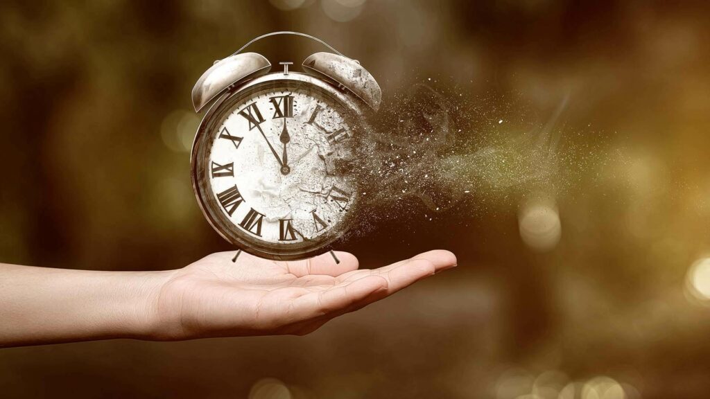 The image features an outstretched human hand holding an antique-style alarm clock. The clock is disintegrating into numerous small particles on one side, signifying the passage of time or the concept of time slipping away. The background is blurred with warm, bokeh light effects that give a sense of a fleeting moment or memory. The overall composition conveys a metaphorical message about the transient nature of time and the importance of cherishing every moment.