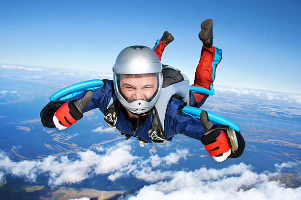 A skydiver in mid-air with a silver helmet and blue jumpsuit smiles and gives thumbs-up while freefalling through a clear blue sky, with a view of clouds and land below.