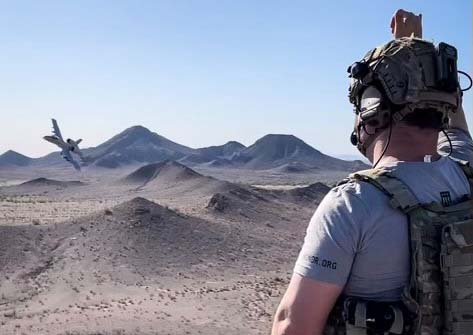 Ramiro Villalobos standing on a desert hillside wearing military communications equipment waving to a fighetr ject passing through the valley below.