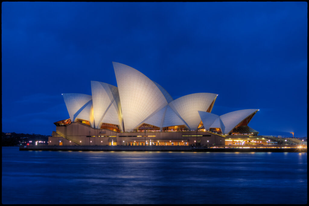 The image depicts the Sydney Opera House, an iconic architectural masterpiece located in Sydney, Australia. The photo showcases the building's distinct sail-like design illuminated against a deep blue evening sky, with its reflection shimmering on the water. The Opera House is renowned for its unique and innovative architecture and is a symbol of Australia.